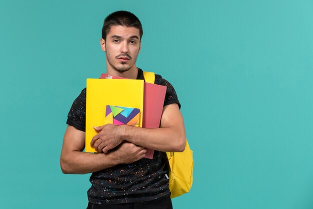 Vista frontal del estudiante en camiseta oscura mochila amarilla con archivos y cuaderno en la pared azul claro