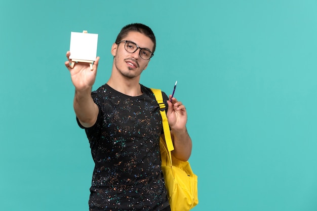 Vista frontal del estudiante en camiseta oscura mochila amarilla con borla y caballete en la pared azul claro