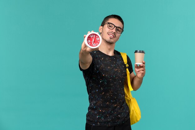 Vista frontal del estudiante en camiseta oscura mochila amarilla con café y relojes en la pared azul