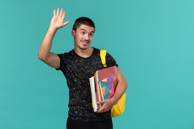 Vista frontal del estudiante en camiseta oscura mochila amarilla con cuaderno y archivos en la pared azul