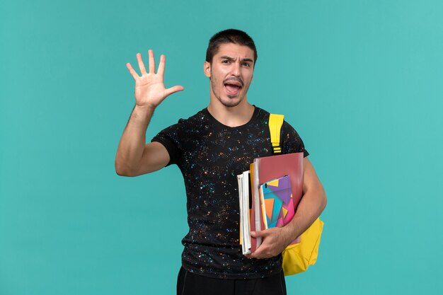 Vista frontal del estudiante en camiseta oscura mochila amarilla con cuaderno y archivos en la pared azul