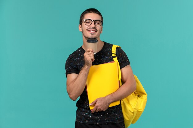 Vista frontal del estudiante en camiseta oscura mochila amarilla con diferentes archivos y café en la pared azul