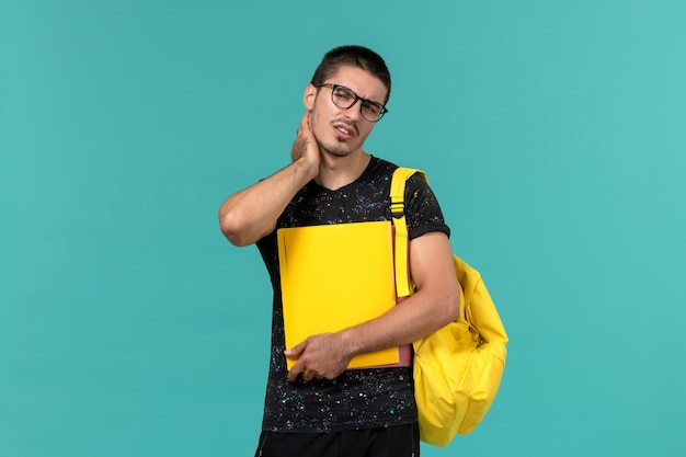 Vista frontal del estudiante en camiseta oscura mochila amarilla con diferentes archivos en la pared azul claro