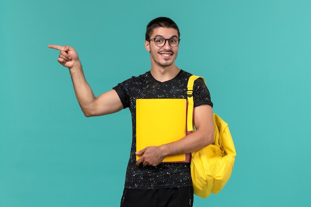 Vista frontal del estudiante en camiseta oscura mochila amarilla con diferentes archivos en la pared azul claro