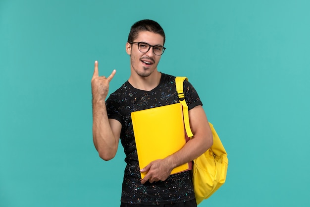 Vista frontal del estudiante en camiseta oscura mochila amarilla con diferentes archivos en la pared azul claro