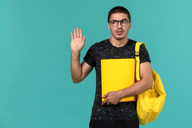 Vista frontal del estudiante en camiseta oscura mochila amarilla con diferentes archivos en la pared azul claro