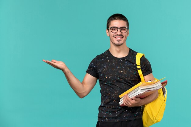 Vista frontal del estudiante en camiseta oscura mochila amarilla sosteniendo archivos y libros sonriendo en la pared azul claro
