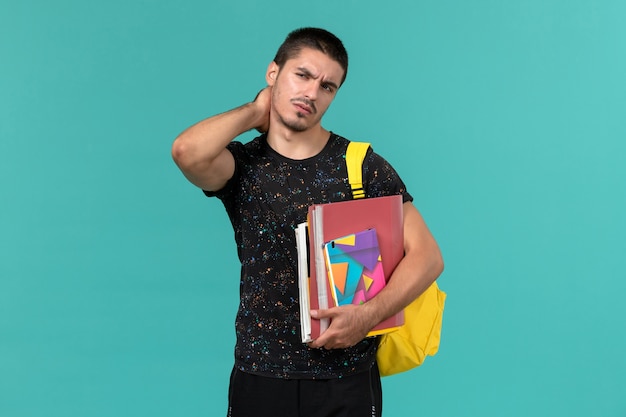 Vista frontal del estudiante en camiseta oscura con mochila amarilla sosteniendo cuaderno y archivos con dolor de cuello en la pared azul