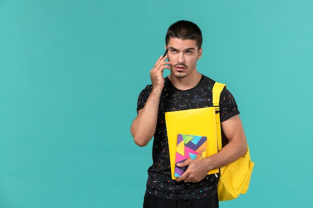 Vista frontal del estudiante en camiseta oscura mochila amarilla sosteniendo un cuaderno y archivos en la pared azul claro