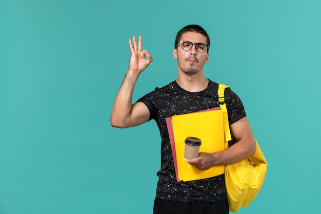 Vista frontal del estudiante en camiseta oscura mochila amarilla sosteniendo diferentes archivos y café en la pared azul