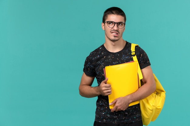 Foto gratuita vista frontal del estudiante en camiseta oscura mochila amarilla sosteniendo diferentes archivos pensando en la pared azul claro