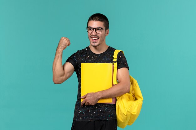 Vista frontal del estudiante en camiseta oscura mochila amarilla sosteniendo diferentes archivos regocijándose en la pared azul claro