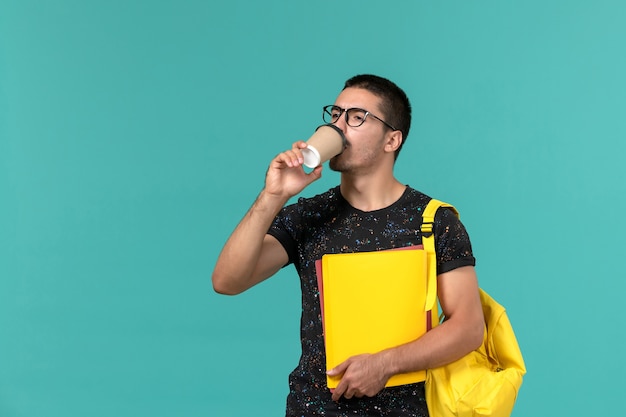 Vista frontal del estudiante en camiseta oscura mochila amarilla sosteniendo diferentes archivos y tomando café en la pared azul