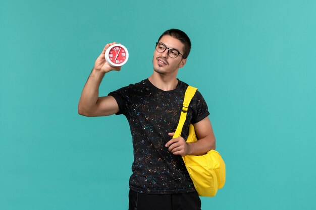 Vista frontal del estudiante en camiseta oscura mochila amarilla sosteniendo relojes en la pared azul