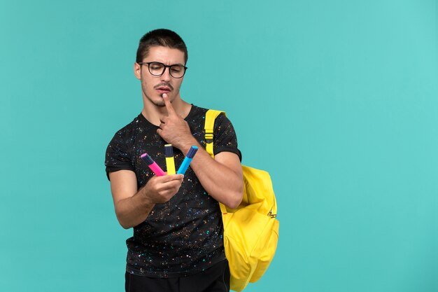 Vista frontal del estudiante en camiseta oscura mochila amarilla sosteniendo rotuladores de colores en la pared azul