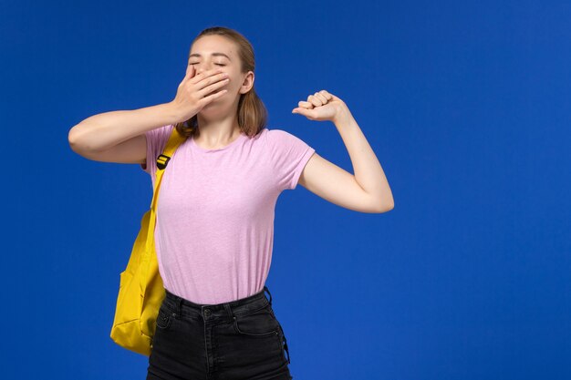 Vista frontal de la estudiante en camiseta rosa con mochila amarilla bostezando en la pared azul claro