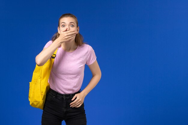 Vista frontal de la estudiante en camiseta rosa con mochila amarilla cerrando la boca en la pared azul claro