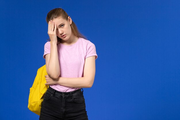 Vista frontal de la estudiante en camiseta rosa con mochila amarilla estresada en la pared azul