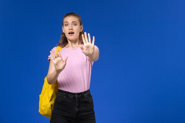 Vista frontal de la estudiante en camiseta rosa con mochila amarilla expresión de miedo en la pared azul claro