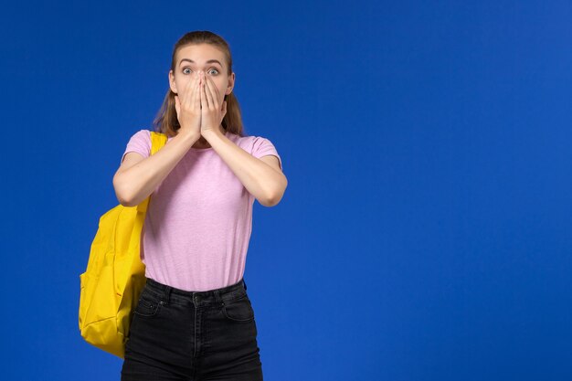 Vista frontal de la estudiante en camiseta rosa con mochila amarilla expresión de sorpresa en la pared azul claro