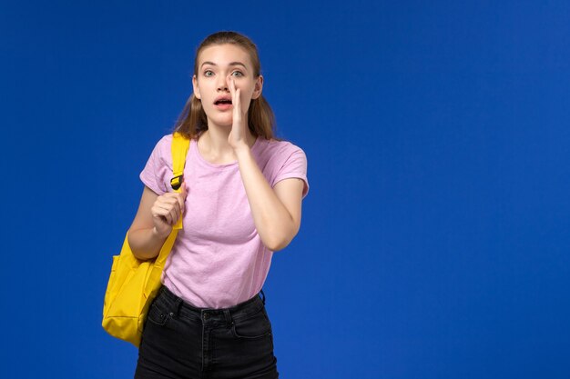 Vista frontal de la estudiante en camiseta rosa con mochila amarilla gritando en la pared azul