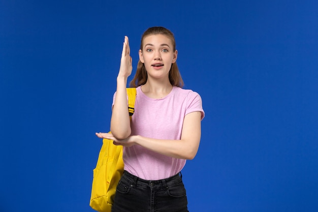 Vista frontal de la estudiante en camiseta rosa con mochila amarilla levantando la mano sobre la pared azul