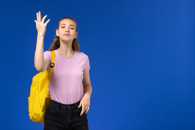 Vista frontal de la estudiante en camiseta rosa con mochila amarilla en la pared azul claro