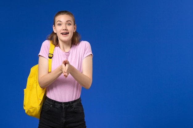 Vista frontal de la estudiante en camiseta rosa con mochila amarilla en la pared azul claro