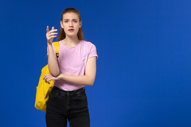 Vista frontal de la estudiante en camiseta rosa con mochila amarilla en la pared azul