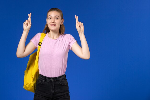 Vista frontal de la estudiante en camiseta rosa con mochila amarilla en la pared azul