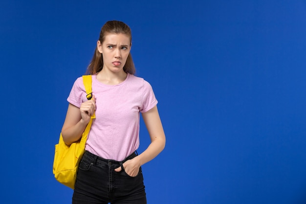 Vista frontal de la estudiante en camiseta rosa con mochila amarilla en la pared azul