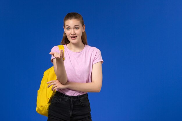 Vista frontal de la estudiante en camiseta rosa con mochila amarilla en la pared azul