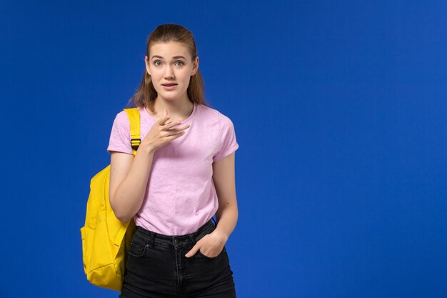 Vista frontal de la estudiante en camiseta rosa con mochila amarilla en la pared azul