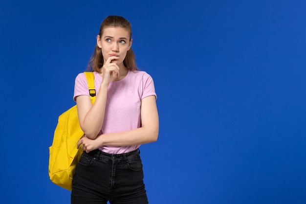 Vista frontal de la estudiante en camiseta rosa con mochila amarilla pensando en la pared azul claro