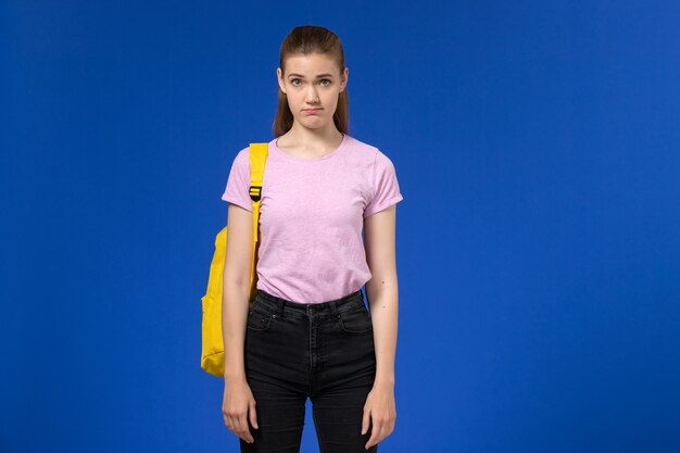 Vista frontal de la estudiante en camiseta rosa con mochila amarilla de pie en la pared azul