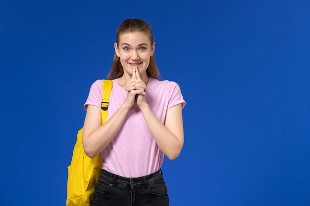 Vista frontal de la estudiante en camiseta rosa con mochila amarilla de pie en la pared azul