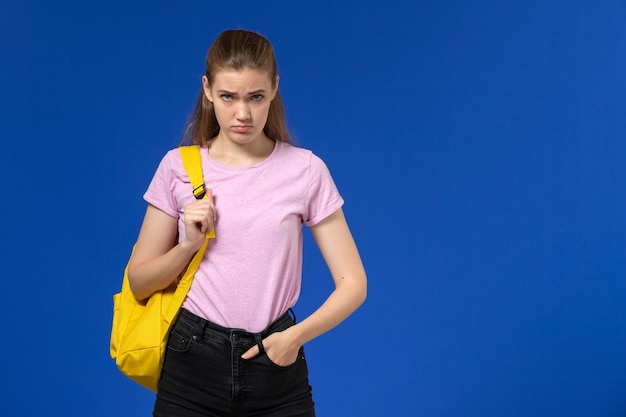 Vista frontal de la estudiante en camiseta rosa con mochila amarilla posando con expresión enojada en la pared azul