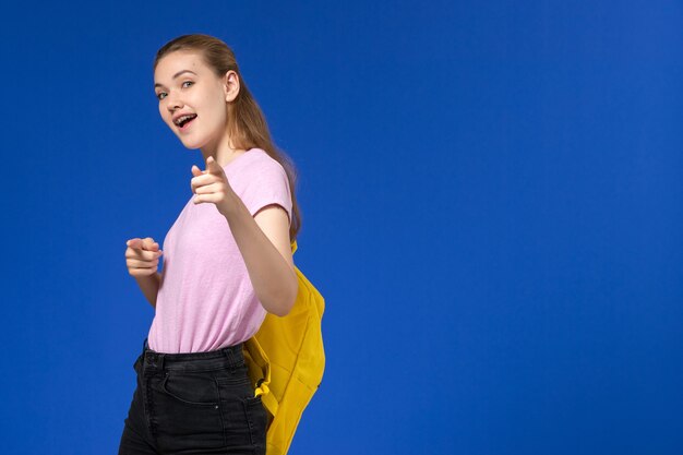 Vista frontal de la estudiante en camiseta rosa con mochila amarilla posando en la pared azul claro