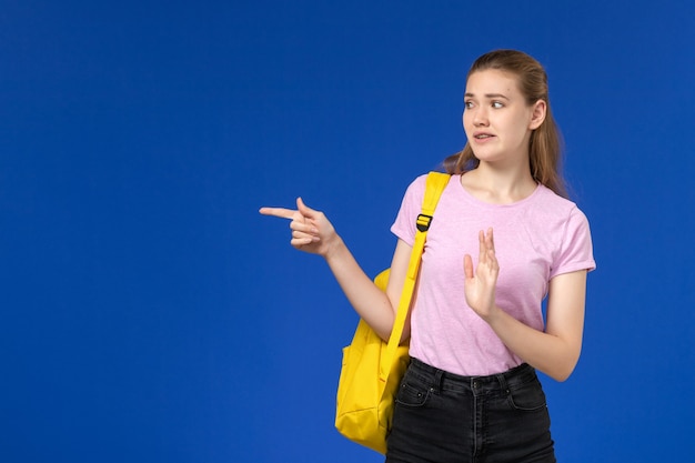 Vista frontal de la estudiante en camiseta rosa con mochila amarilla posando en la pared azul claro