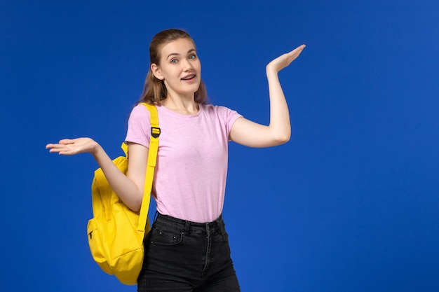 Vista frontal de la estudiante en camiseta rosa con mochila amarilla posando en la pared azul claro