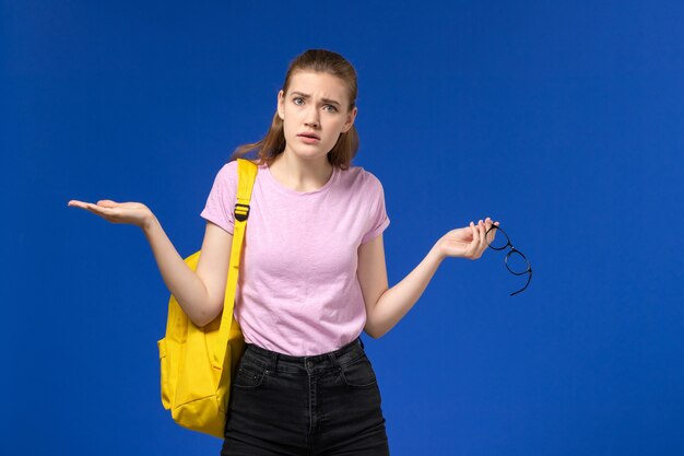 Vista frontal de la estudiante en camiseta rosa con mochila amarilla posando en la pared azul