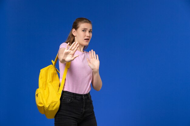 Vista frontal de la estudiante en camiseta rosa con mochila amarilla posando en la pared azul