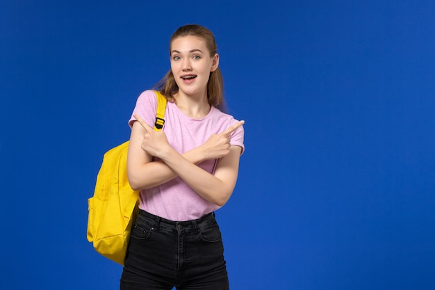 Vista frontal de la estudiante en camiseta rosa con mochila amarilla posando en la pared azul