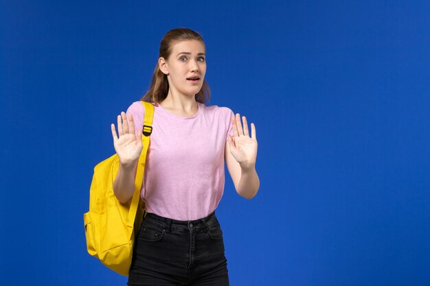 Vista frontal de la estudiante en camiseta rosa con mochila amarilla posando en la pared azul