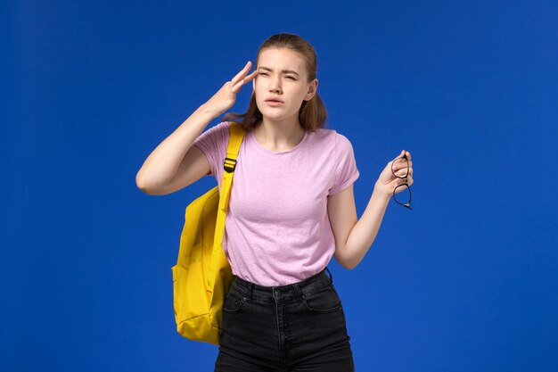 Vista frontal de la estudiante en camiseta rosa con mochila amarilla posando en la pared azul