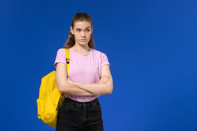 Vista frontal de la estudiante en camiseta rosa con mochila amarilla posando en la pared azul