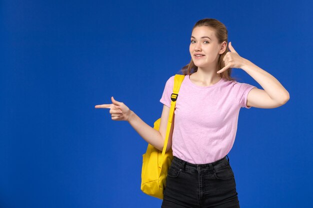 Vista frontal de la estudiante en camiseta rosa con mochila amarilla posando sonriendo en la pared azul claro