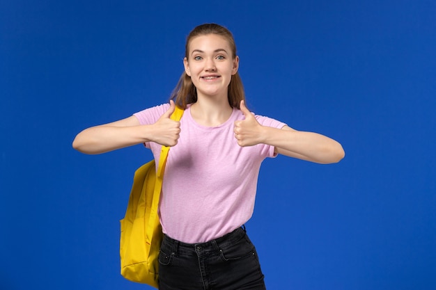 Vista frontal de la estudiante en camiseta rosa con mochila amarilla posando y sonriendo en la pared azul