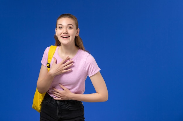 Vista frontal de la estudiante en camiseta rosa con mochila amarilla riendo en la pared azul