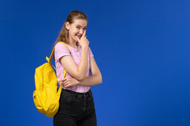 Vista frontal de la estudiante en camiseta rosa con mochila amarilla riendo en la pared azul